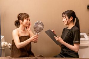 Beauty Salon Lilyでカウンセリングを受ける女性。鏡で自分の顔を確認し、エステティシャンがタブレットに記録している